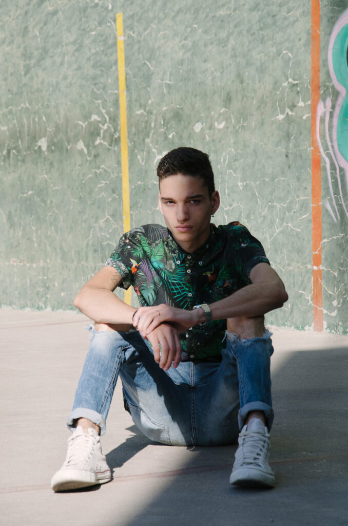 Victor Zepeda sits against a graffiti-covered wall wearing a tropical shirt and blue jeans with a watch on his wrist.